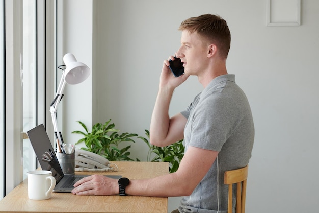 Young man sitting at desk in front of window and taking on phone