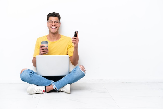 Young man sitting on the floor isolated on white background holding coffee to take away and a mobile