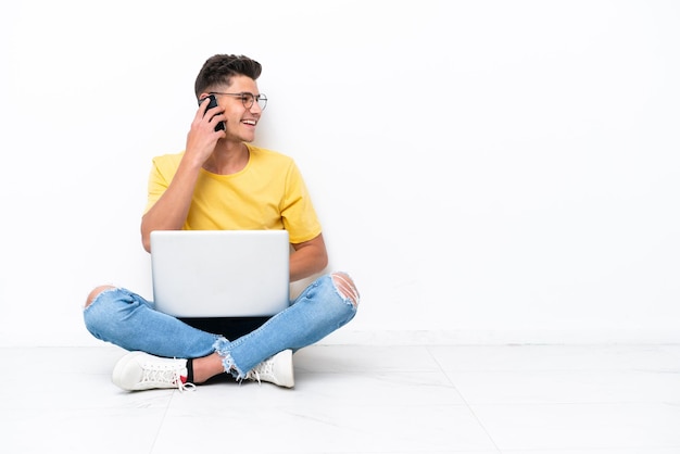 Young man sitting on the floor isolated on white background keeping a conversation with the mobile phone with someone