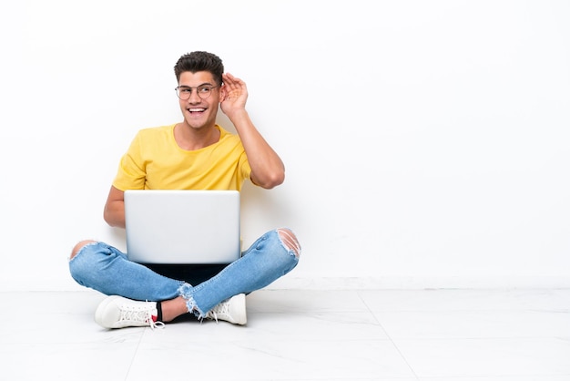 Young man sitting on the floor isolated on white background listening to something by putting hand on the ear