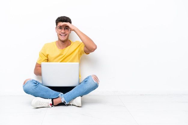 Young man sitting on the floor isolated on white background looking far away with hand to look something