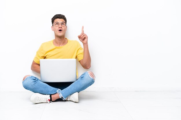 Young man sitting on the floor isolated on white background pointing up and surprised