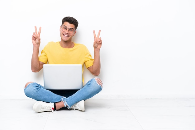 Young man sitting on the floor isolated on white background showing victory sign with both hands