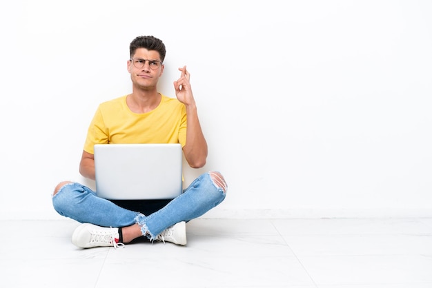 Young man sitting on the floor isolated on white background with fingers crossing and wishing the best