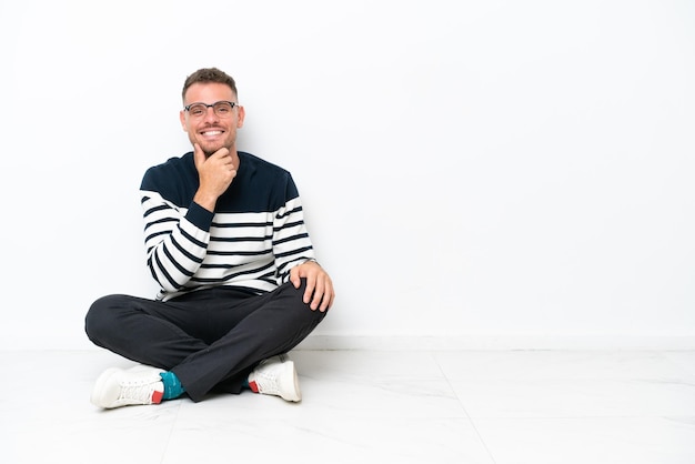 Young man sitting on the floor isolated on white background with glasses and smiling