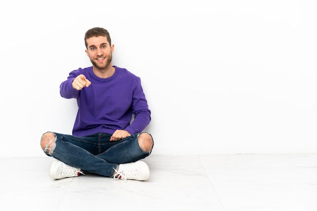 Young man sitting on the floor surprised and pointing front