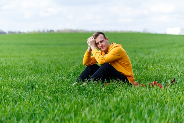 Photo a young man sitting on green grass is thinking about the future. handsome guy is relaxing in the park on a sunny summer day. vacation concept