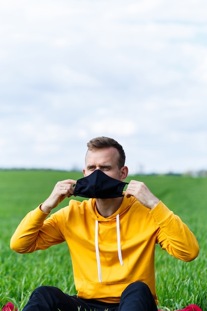 Young man sitting on green grass in a protective antiviral mask and thinking about the future. The guy is resting in the park on a sunny summer day. The concept of rest. Virus protection. Quarantine