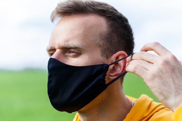 Young man sitting on green grass in a protective antiviral mask and thinking about the future. The guy is resting in the park on a sunny summer day. The concept of rest. Virus protection. Quarantine