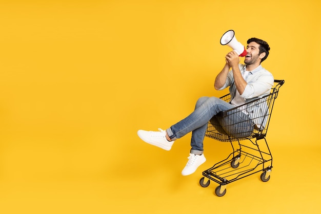 Young man sitting inside of shopping trolley and holding megaphone  isolated on yellow background