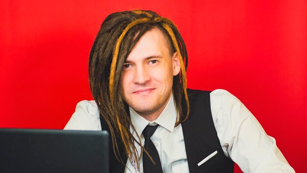 Young man sitting at laptop looking at camera and smiling on red background Positive freelancer works remotely at modern workplace