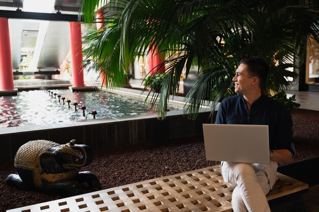 Young man sitting in a public place looking the side smiling while using computer laptop