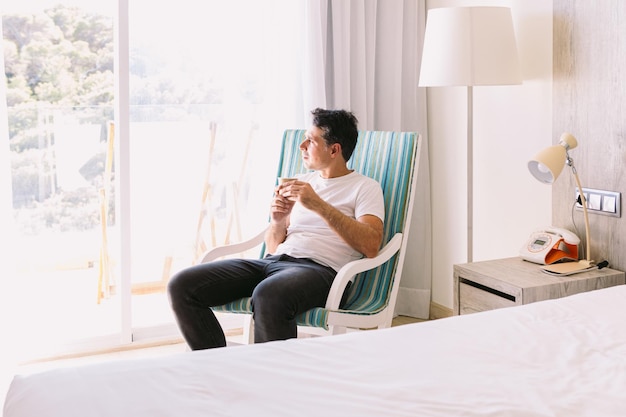 Young man sitting in a rocking chair in his bedroom having a drink with the light coming through the window Vacation relaxation rest breakfast coffee tea and hotel concept