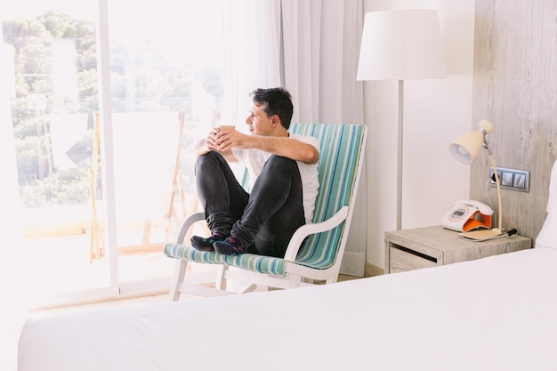 Young man sitting in a rocking chair in his bedroom having a drink with the light coming through the window Vacation relaxation rest breakfast coffee tea and hotel concept