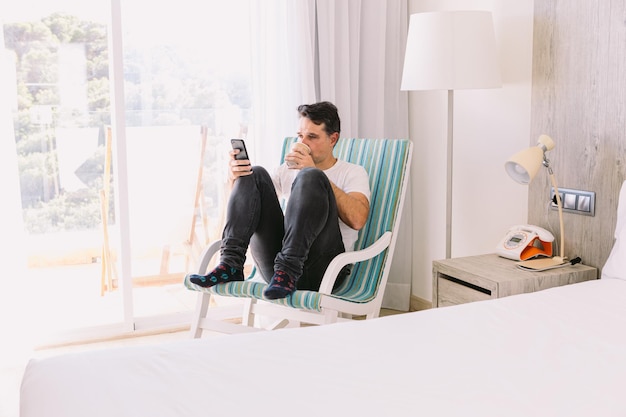 Young man sitting in a rocking chair in his bedroom looking at his cell phone and having a coffee with the light coming through the window Concept of working vacation connection and smartphone