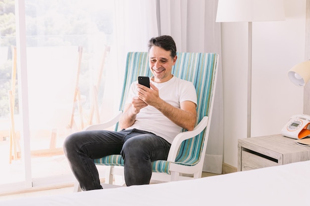 Young man sitting in a rocking chair in his bedroom with his legs crossed looking at his cell phone with light coming through the window Concept of working vacation connection and smartphone