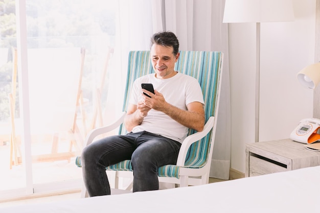 Young man sitting in a rocking chair in his bedroom with his legs crossed looking at his cell phone with light coming through the window Concept of working vacation connection and smartphone