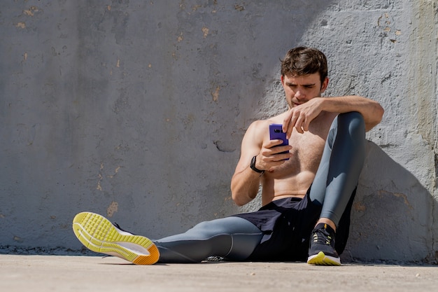 Young man sitting shirtless using mobile and rested after training