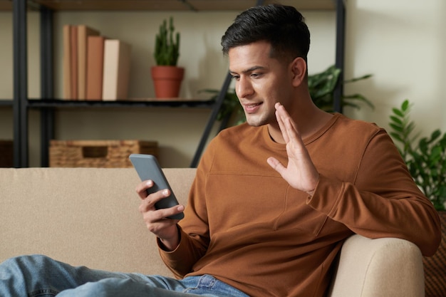 Young man sitting on sofa in the living room and waving his hand to camera of his mobile phone during online conversation