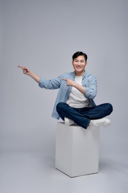 A young man sitting on white cube pointing with his hands to the side