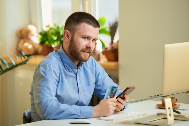 A young man sitting at a white desk in front of a laptop and holds a smartphone and a credit card in his hands at home