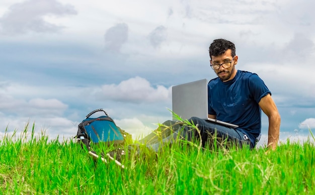 Young man sitting with laptop and backpack in the field Man sitting in the green field working from his laptop Freelancer man concept working from the field