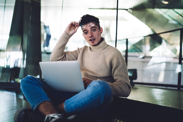 Young man sitting with laptop on knees and raising glasses