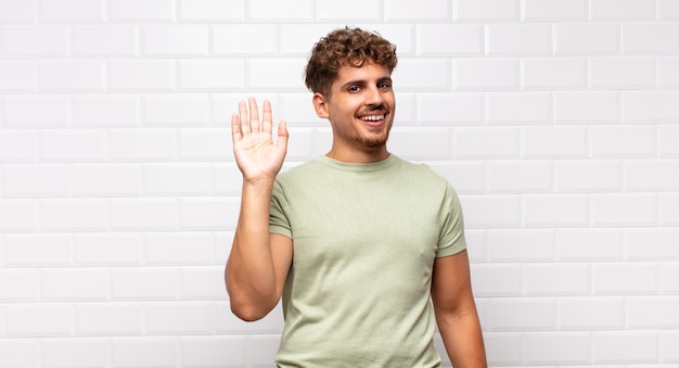 Young man smiling happily and cheerfully, waving hand, welcoming and greeting you, or saying goodbye