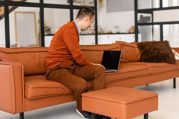 Young man smiling, reads the screen of a laptop computer while relaxing on a comfortable couch at home.