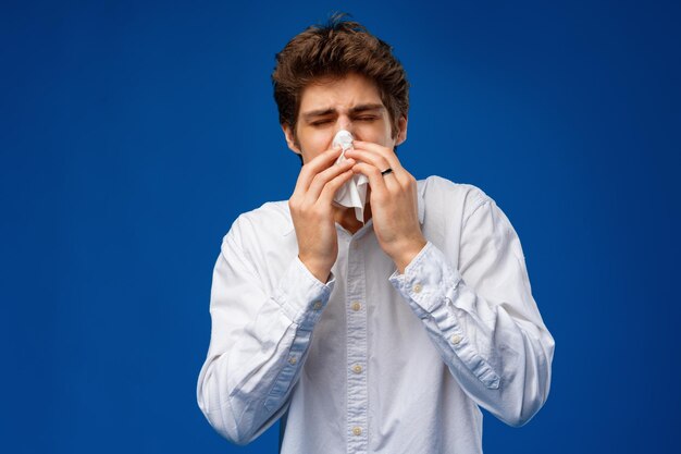 A young man sneezes into a napkin over blue background