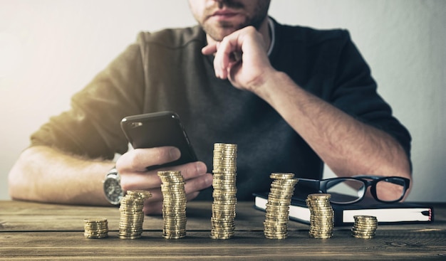 young man behind stacked coins checking his Finances in a smartphone to save his money. ideal for websites and magazines layouts