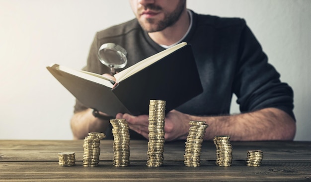 young man behind stacked coins checking with magnifying glass his Finances in a book to save his money. ideal for websites and magazines layouts