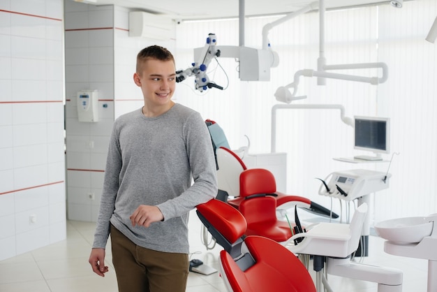 A young man stands near a red dental chair and smiles in modern white dentistry Treatment and prevention of caries from youth Modern dentistry and prosthetics