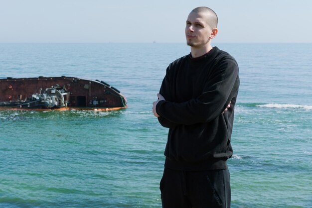 A young man stands with crossed hands on the coastline and looks sideway with a sunken ship in the background Male Man Sea Caucasian Portrait Nature Face Young Adult Water Beach Summer