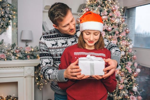 Young man stands behind woman. They hold box with present together. He looks at her. She looks at present. They are in decorated room.