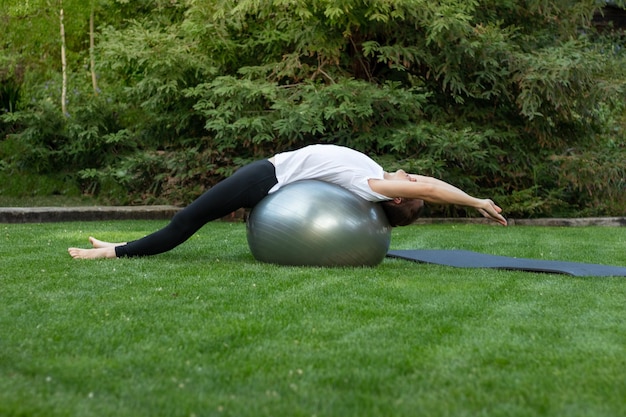 Young man stretching his hole body on a pilates ball to improve his back pain in the garden for better brething