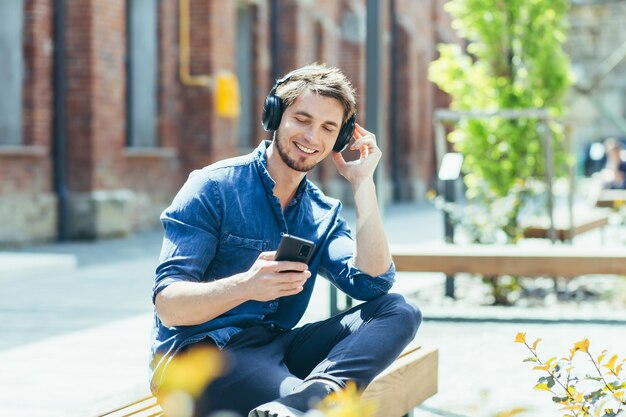 Photo young man student guy sitting on campus on a bench in the lotus position listening to an audiobook audio lecture in headphones holding a phone in his hands he smiles