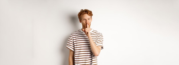 Photo young man student with red hair shushing telling a secret and smiling standing over white              person