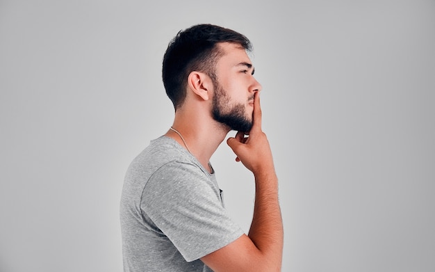 A young man in the studio thinks on a gray background, putting a finger on his lips