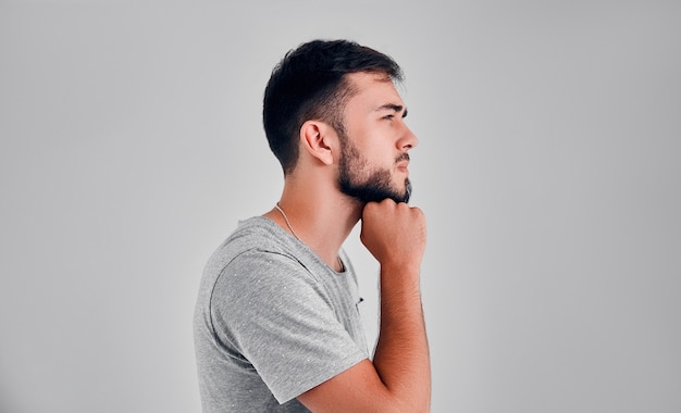 A young man in the studio thinks on a gray background, putting a fist under his chin