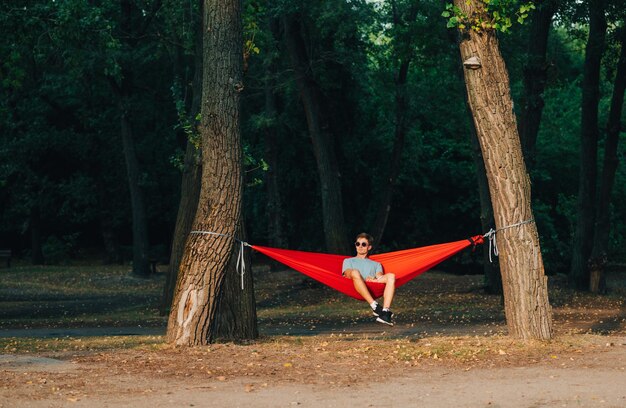 Photo young man in stylish casual clothes and sunglasses sitting in a hammock on a background of the park and admiring the sunset relax outdoors