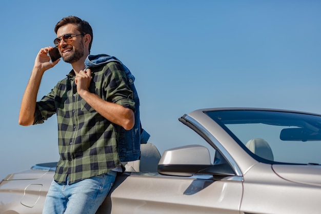 Young man in sunglasses standing at the car and having conversation on the phone