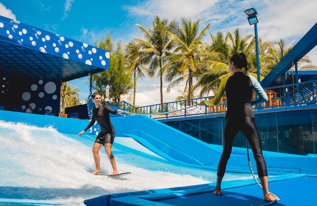 Young man surfing with trainer on a wave simulator at a water amusement park