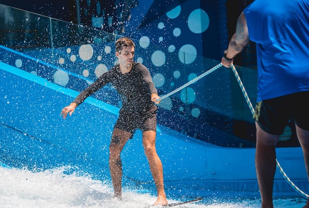 Young man surfing with trainer on a wave simulator at a water amusement park