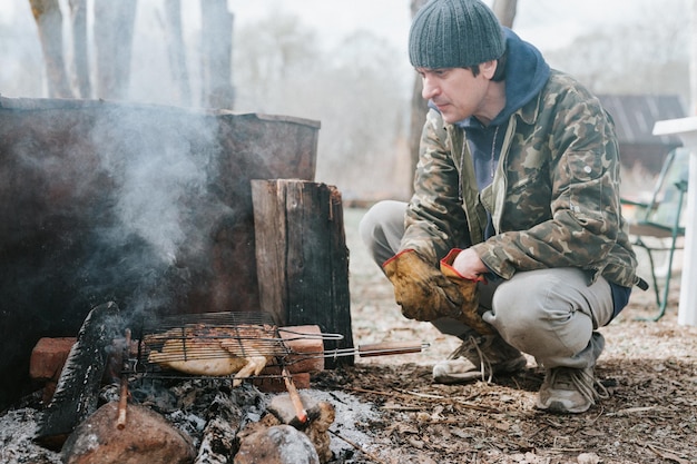 Young man survivalist cooks roasts chicken meat food are fried on grill on smoldering coals or ember from a campfire on the ground barbecue in camping conditions countryside and wild rustic life