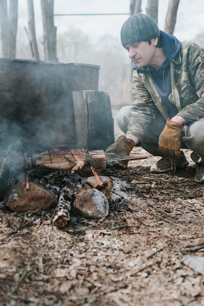 Young man survivalist cooks roasts chicken meat food are fried on grill on smoldering coals or ember from a campfire on the ground barbecue in camping conditions countryside and wild rustic life