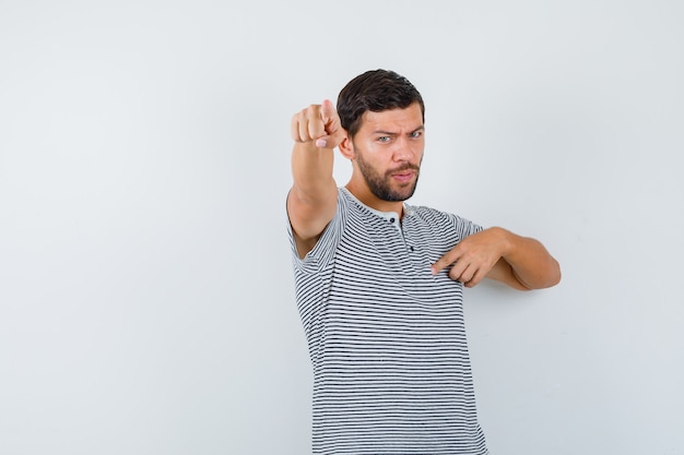 Young man in t-shirt pointing forward and at himself and looking indecisive , front view.