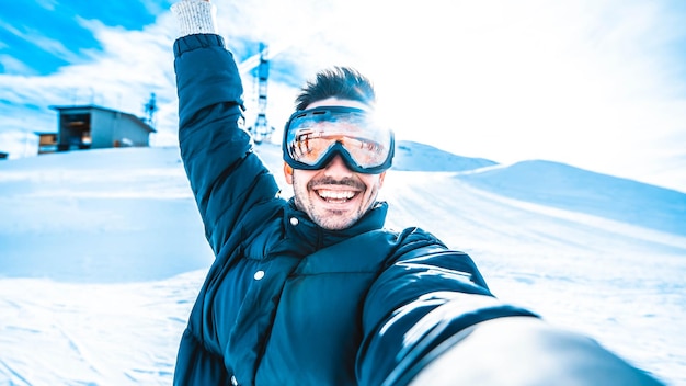 Young man taking selfie picture with smart cell phone hiking mountain on snowy slope - Happy skier having fun in ski resort vacation - Winter lifestyle, extreme sport and happy people concept