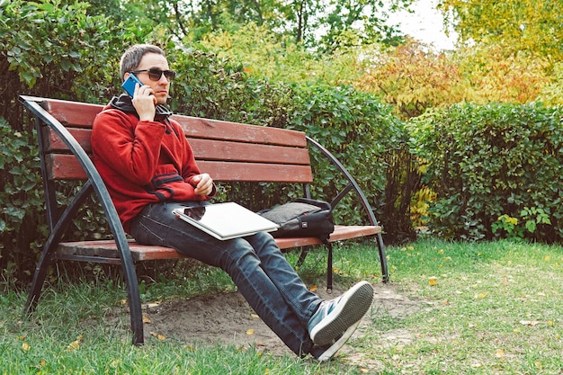 Young man talking on mobile phone during distance work on laptop computer sitting in city park in day Hipster guy calling via cellphone and reading information from ultrabook solving issues