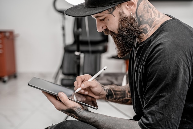 Young man tattoo artist with beard holding pencil and sketch looking positive and happy standing and smiling in workshop place.